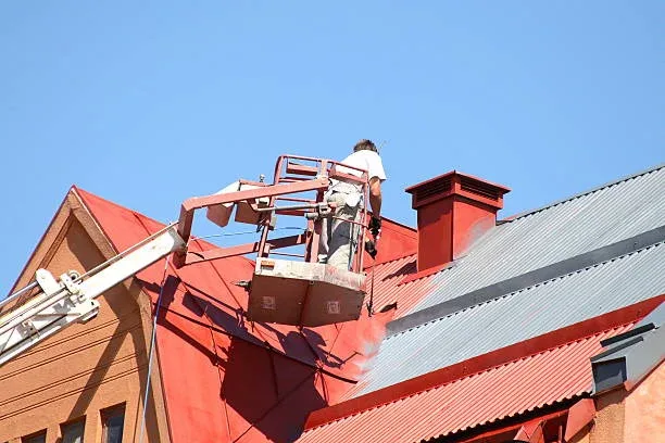 Painter working from a boom lift while spray-coating a red metal roof in Hamilton